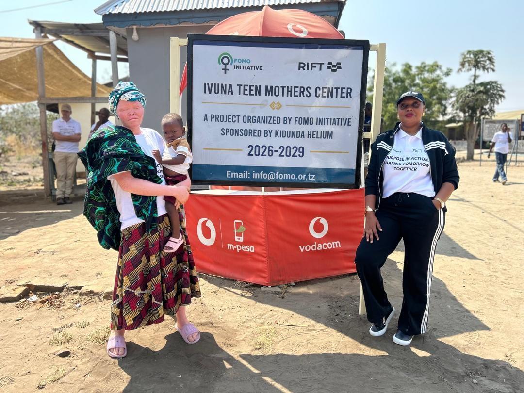 A programme participant holding her child and Vivian Sirikwa beside the Ivuna Teen Mothers Centre signboard